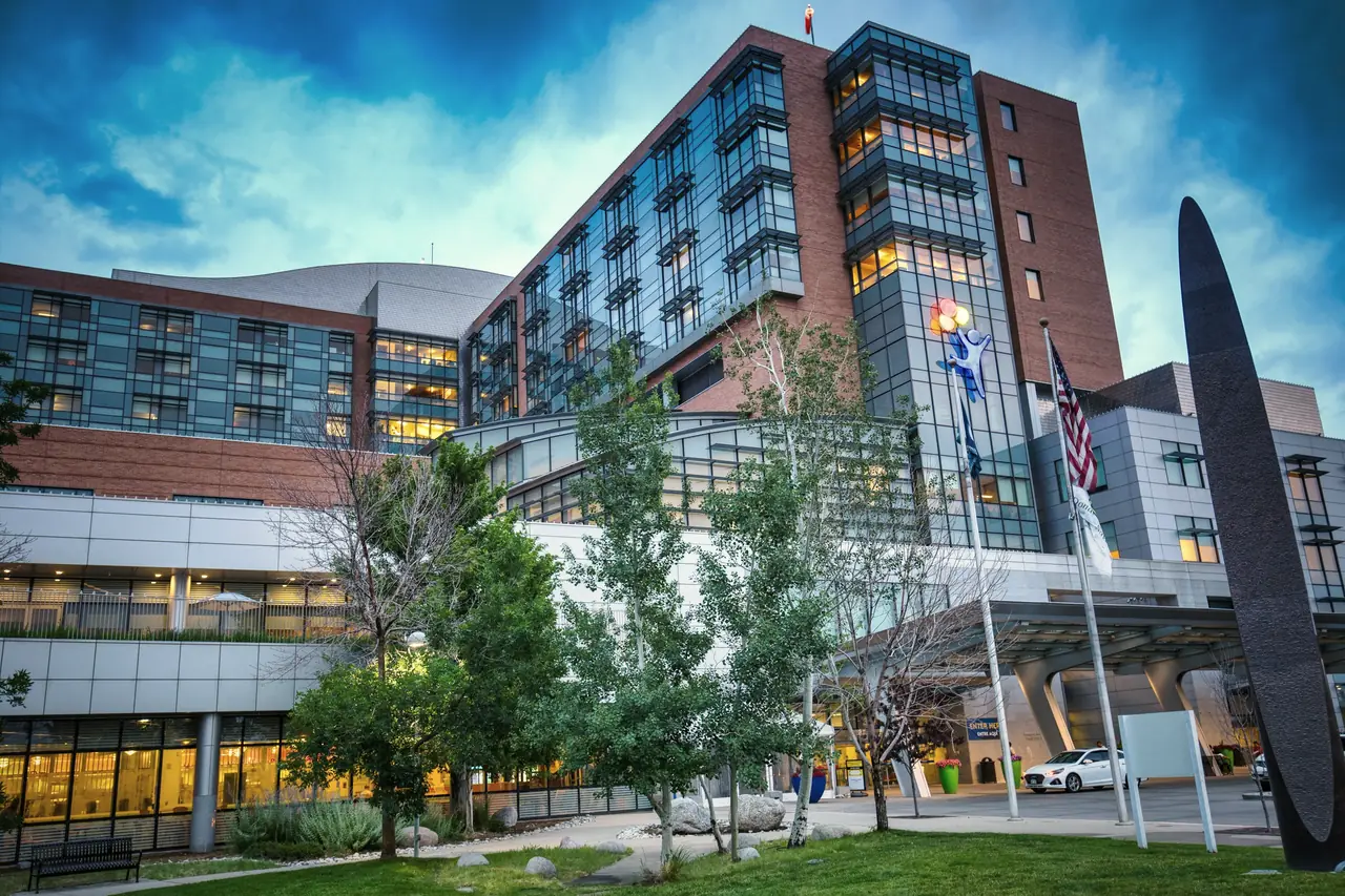 A hospital campus at dusk, the ground-floor glazing lit from inside and the patient tower rising behind it.